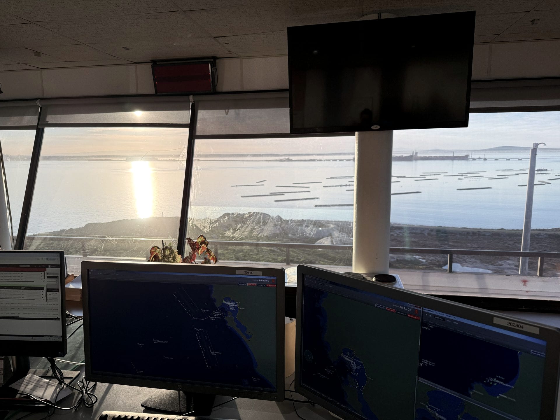 Operations vantage over Saldanha Bay, South Africa at last light: vessel-tracking and electronic-chart monitors with the bay, moored craft and headland beyond, where Afrishore coordinates west-coast port calls